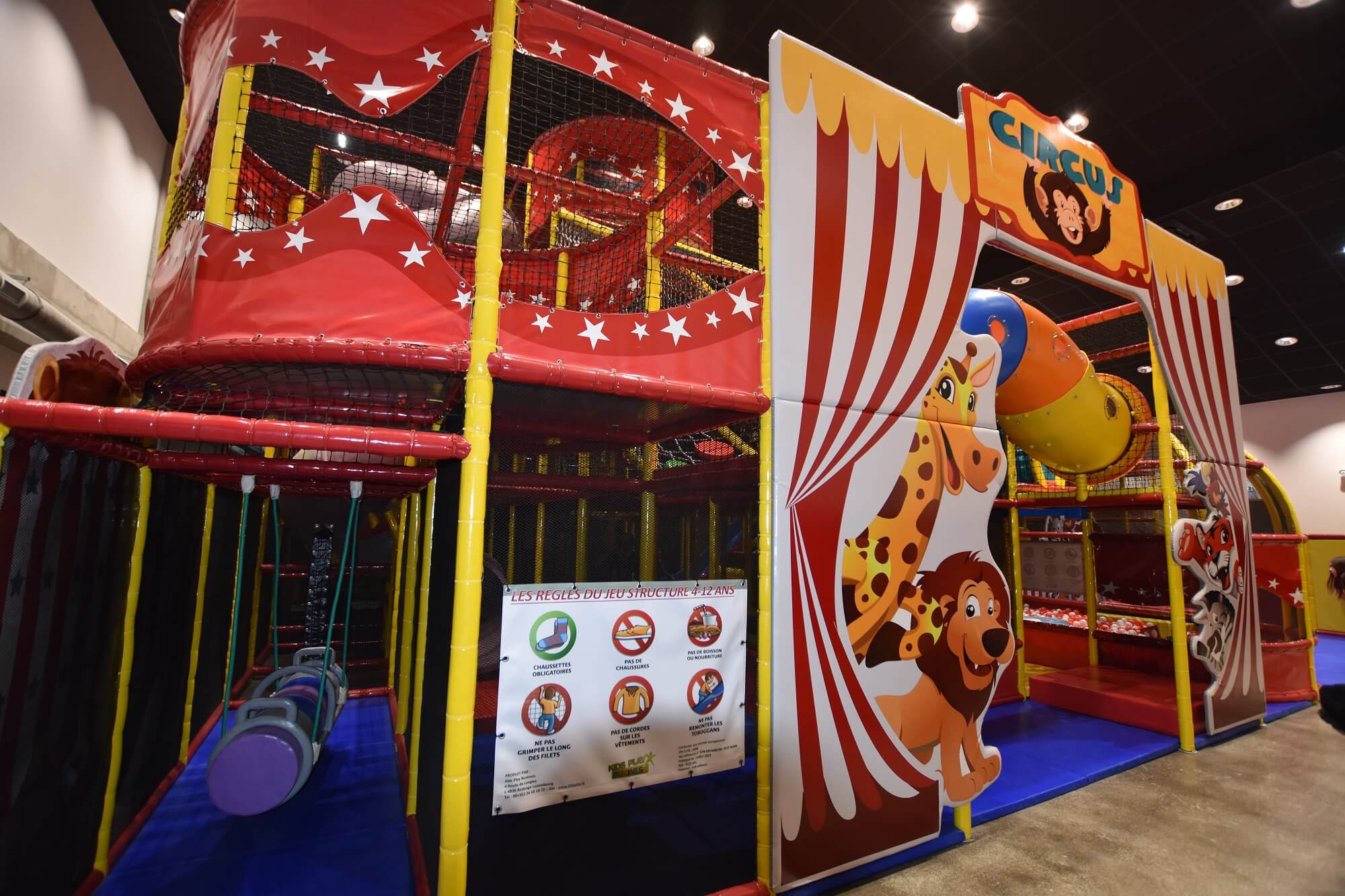 Façade de la structure tubulaire Kids Park sur le thème du cirque au BAX Bowling de Brumath. Réalisation Play In Business montrant l'arche d'entrée décorée d'animaux de la savane, une petite balançoire et le panneau des consignes de sécurité.