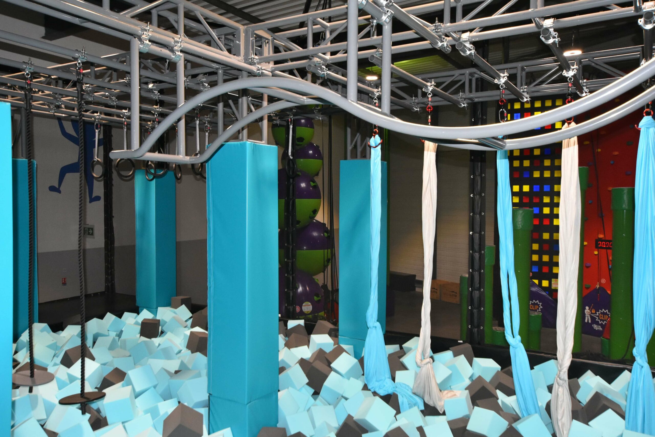 Parcours Ninja suspendu au-dessus d'une grande fosse à cubes de mousse gris et bleus chez Alti Arena au Puy-en-Velay. Aménagement Play In Business comprenant des piliers de protection, des anneaux suspendus et des tissus aériens.