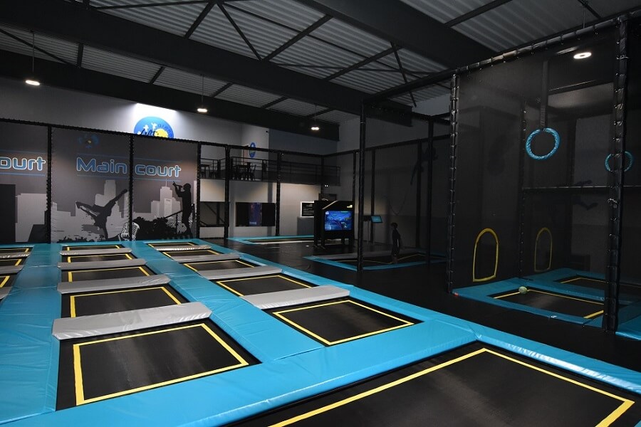 Zone de saut libre (Main Court) du Trampoline Park Alti Arena au Puy-en-Velay. Réalisation Play In Business montrant un ensemble de trampolines noirs et bleus avec bordures jaunes, et un mur interactif en arrière-plan.