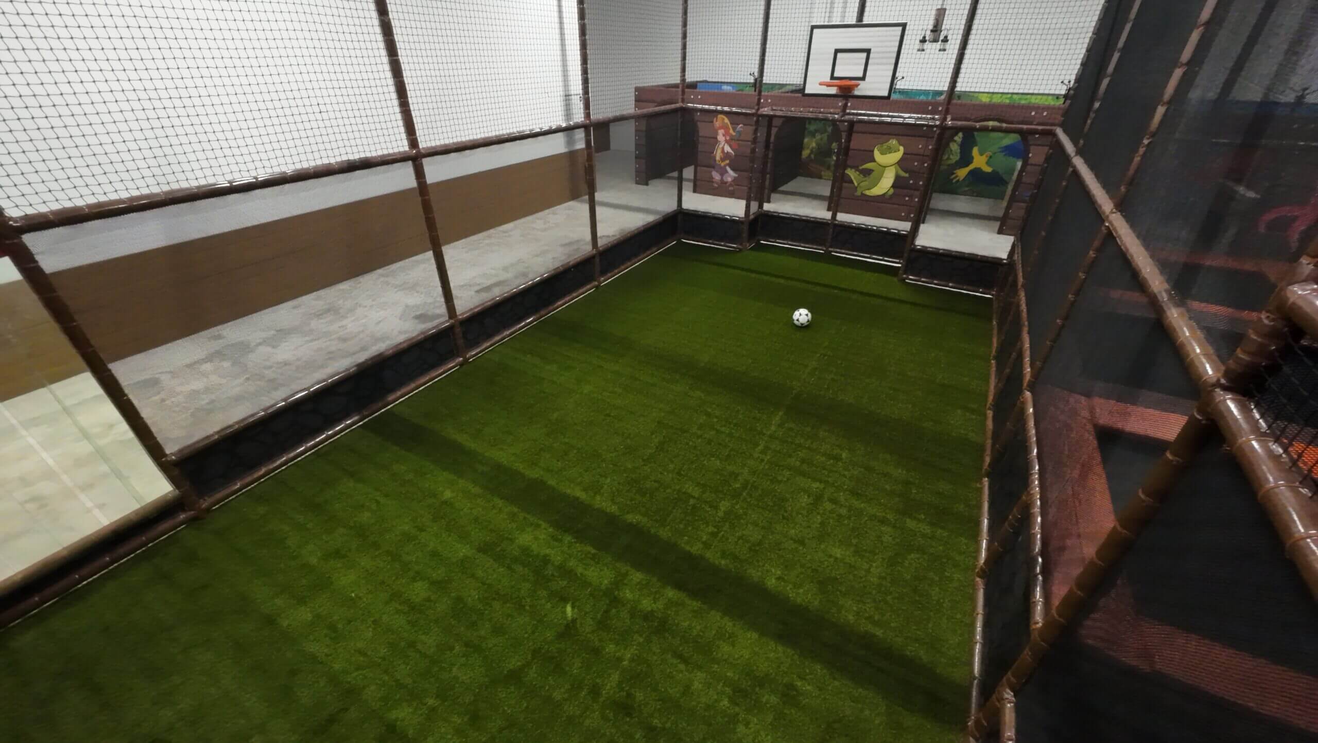 Arène multisport indoor aménagée avec gazon synthétique, panier de basket et zone de football, sécurisée par des filets et mousses de protection pour parc de loisirs couvert.
