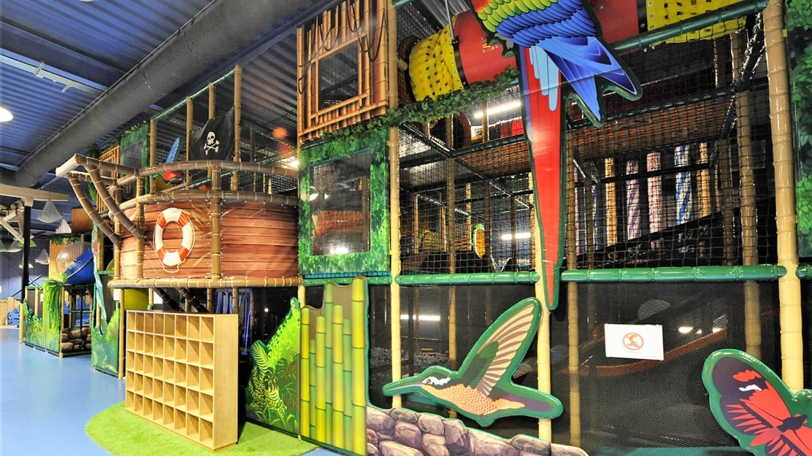 Structure tubulaire géante sur le thème de la jungle et des pirates chez Big Little à Wittenheim. Aménagement Play In Business incluant des décors d'animaux exotiques, un bateau pirate et des rangements à chaussures au premier plan.