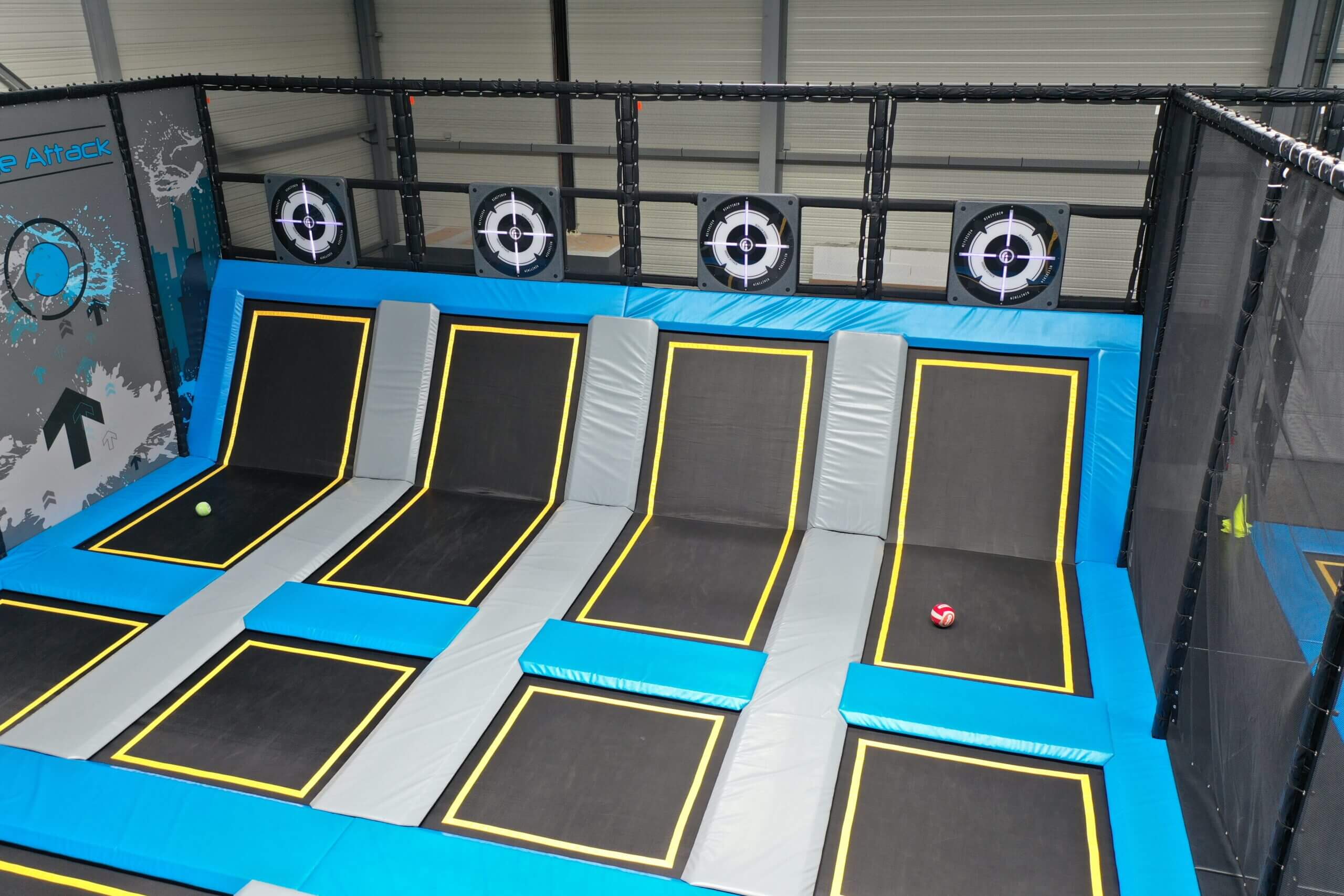 Zone de trampoline park aménagée avec le jeu interactif Dodge Attack, montrant des trampolines bleus et noirs surmontés de cibles lumineuses murales pour parc de loisirs indoor.