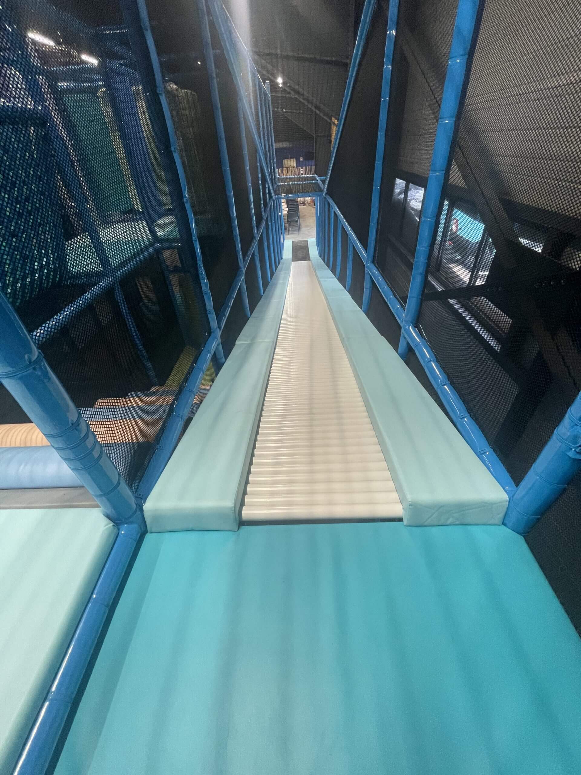 Toboggan à rouleaux 'Roller Run' vu de haut, sécurisé par des mousses bleues et des filets, intégré dans une structure de jeux tubulaire pour parc de loisirs indoor.