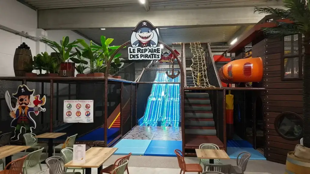 Aire de jeux couverte Le Repaire des Pirates avec des toboggans lumineux bleus, un filet d'escalade, un décor de bateau pirate et un espace restauration au premier plan.
