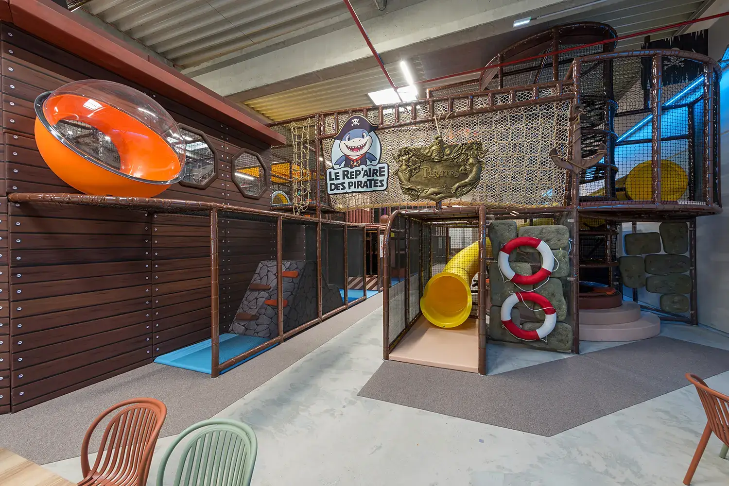 Structure de jeux multiniveaux Le Repaire des Pirates avec un toboggan tube jaune, un parcours sécurisé par des filets, une façade de navire en bois et des décors maritimes.