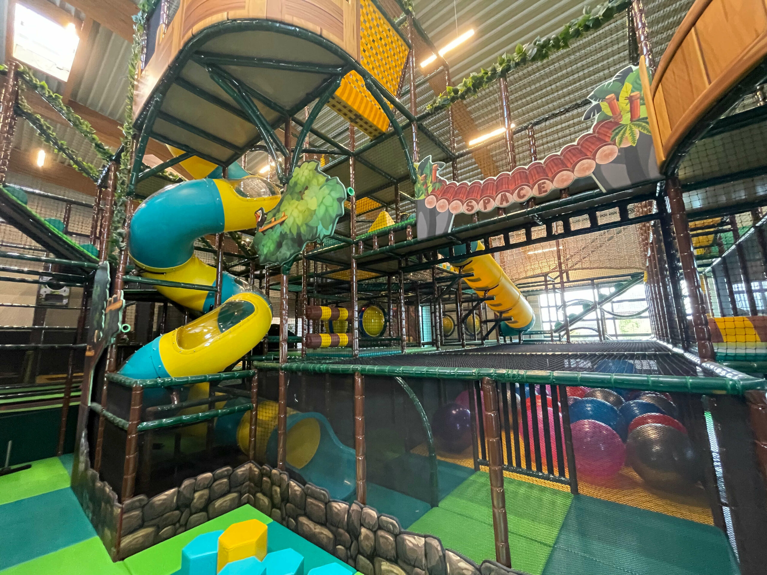 Vue détaillée de l'immense structure de jeux tubulaire sur le thème de la jungle au S'pace Lontzen. Réalisation Play In Business montrant un toboggan en spirale jaune et bleu, des décors de végétation, et des ponts suspendus sécurisés par des filets.