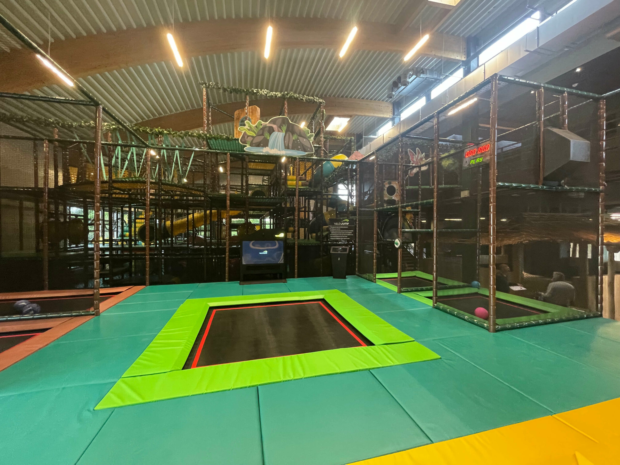 Zone de trampolines avec tapis de protection verts et écran de jeu interactif au Jungle Park du S'pace Lontzen. Aménagement sur mesure par Play In Business avec, en arrière-plan, la grande plaine de jeux thématisée aventure.