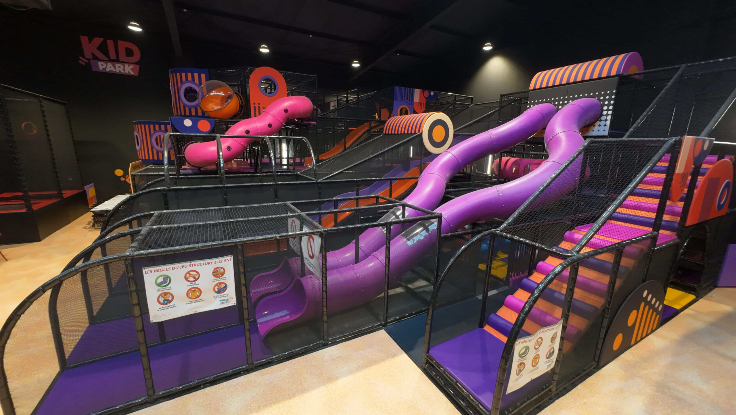 Vue de face de la grande structure de jeux tubulaire du Kid Park au Monky Châteauroux. Installation Play In Business au design moderne, montrant de grands toboggans violets, des escaliers en mousse orange et des filets de sécurité.