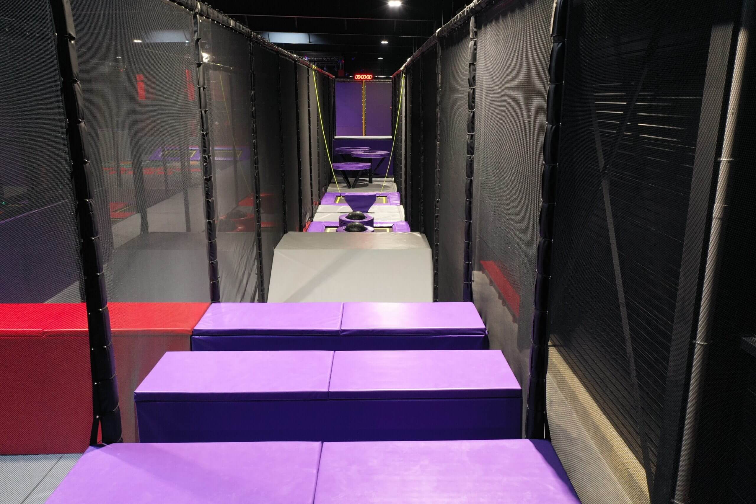 Ligne de départ du Parcours Bounce : une course d'obstacles indoor comprenant des modules d'agilité en mousse violette et des trampolines, entièrement sécurisée par des filets noirs pour trampoline park.