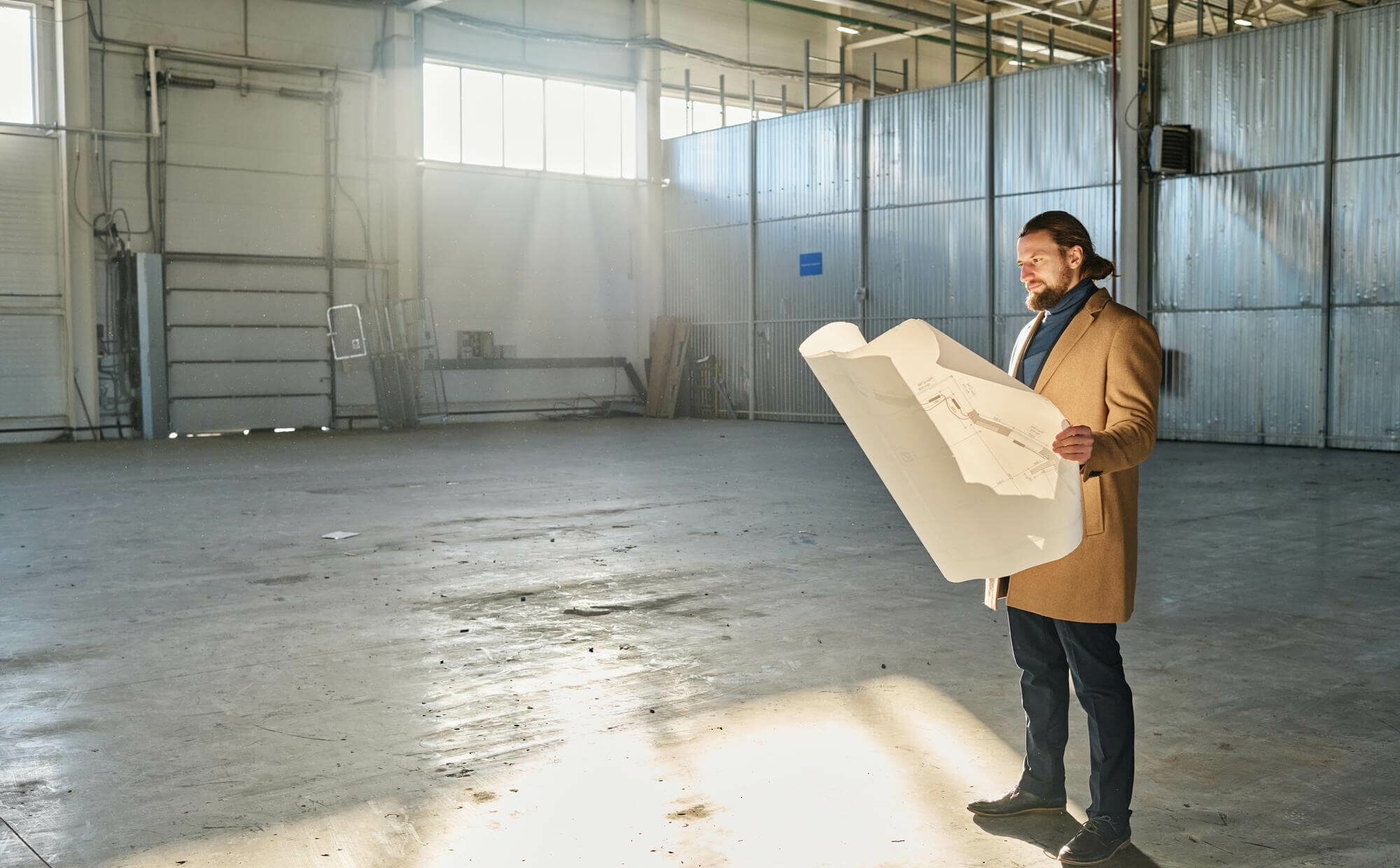 Un entrepreneur en tenue business décontractée tenant un dossier de documents (devis, plans) dans un grand bâtiment brut et lumineux, symbolisant la planification de son futur parc de loisirs.