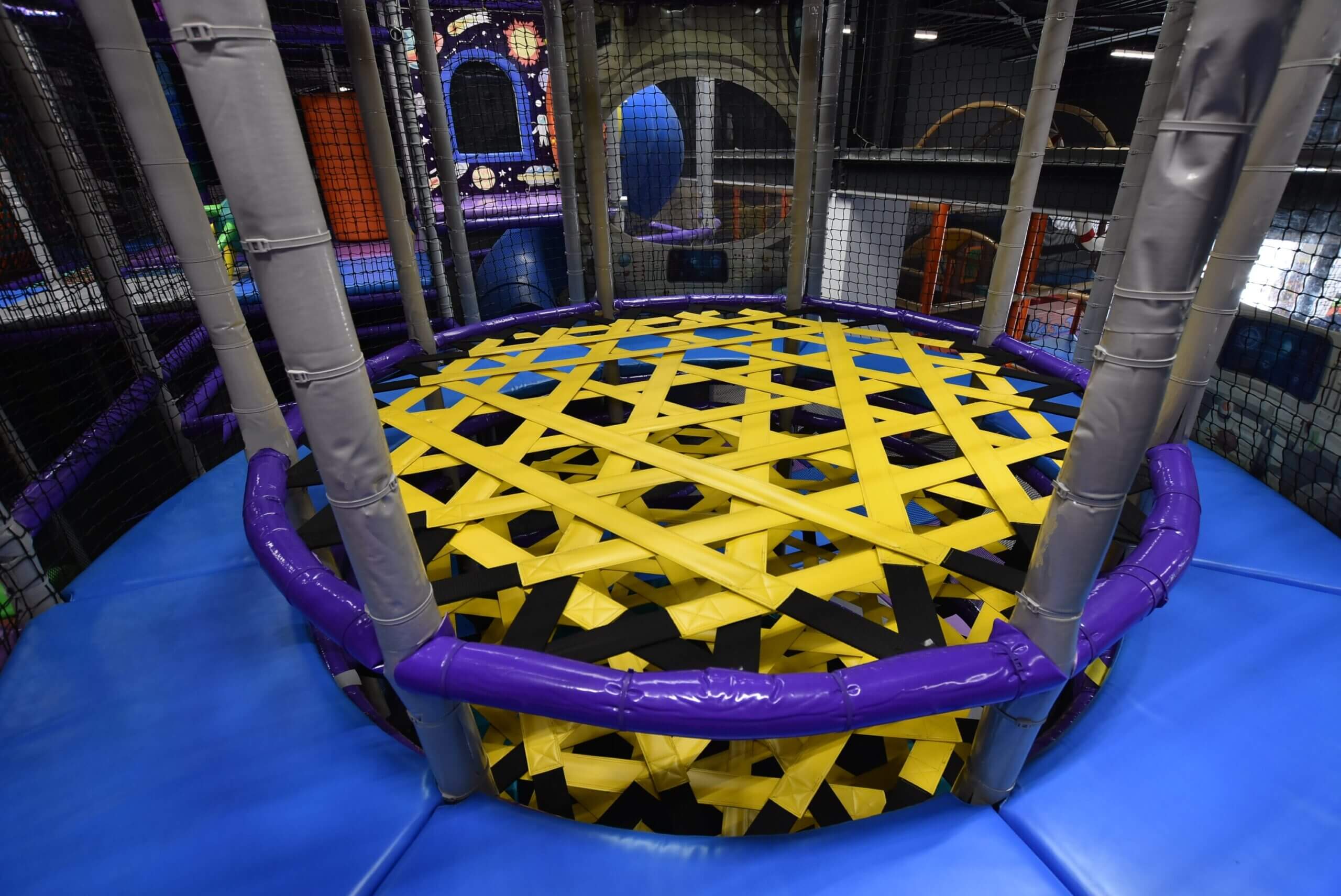 Vue plongeante de la tour à élastiques, un puits de descente circulaire traversé par un réseau tissé d'élastiques jaunes et noirs, sécurisé par un rembourrage de protection violet et bleu dans une structure de jeux intérieure.