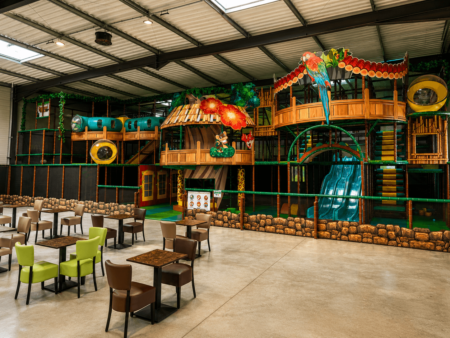 Vue d'ensemble de l'immense plaine de jeux sur le thème de la jungle chez Wistitiparc à Saint-Pierre-lès-Elbeuf. Réalisation Play In Business montrant des décors de perroquet, un toboggan géant bleu et l'espace cafétéria au premier plan.