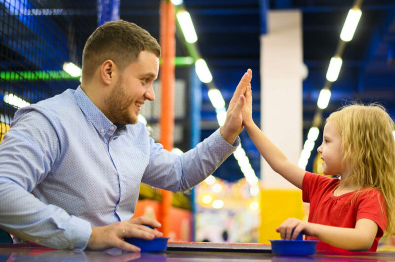 Père et fille jouant au air hockey dans un parc indoor. Play In Business conçoit vos espaces de loisirs en intégrant des activités annexes pour toute la famille.